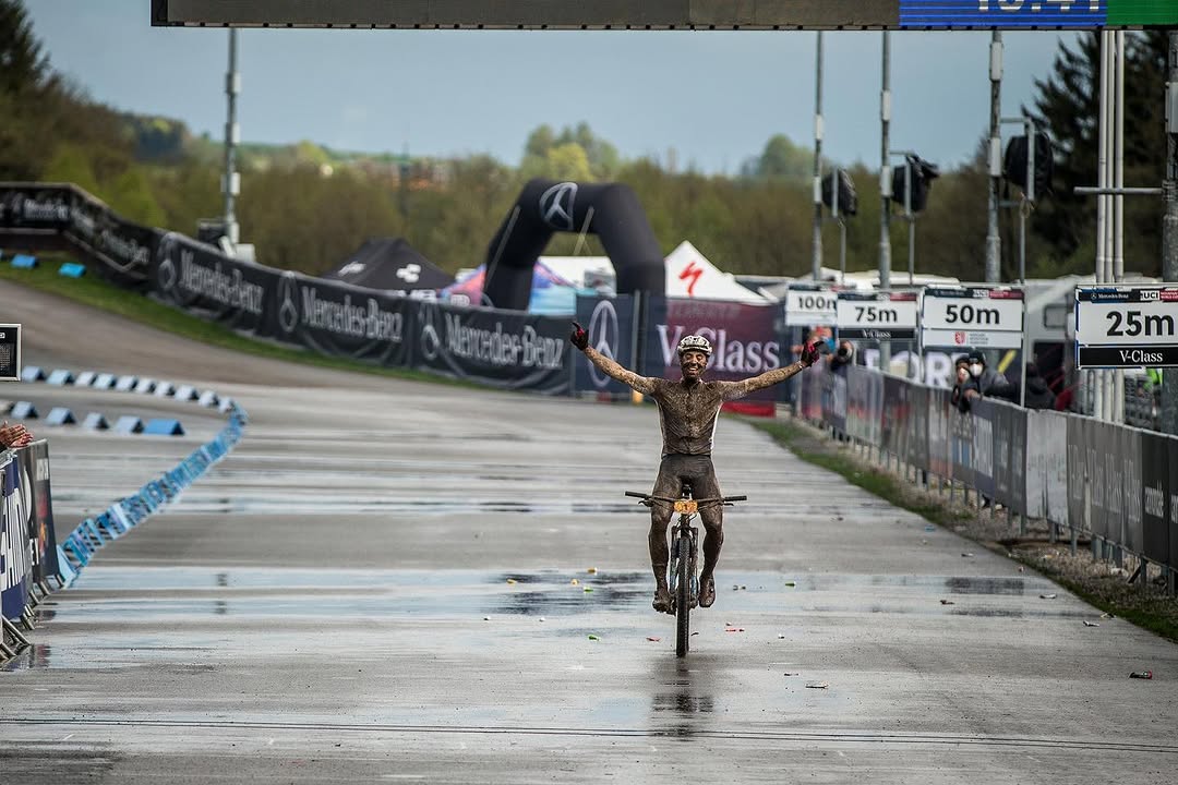 Carter Woods crossing the finish line at Albstadt, arms spread wide, covered in mud